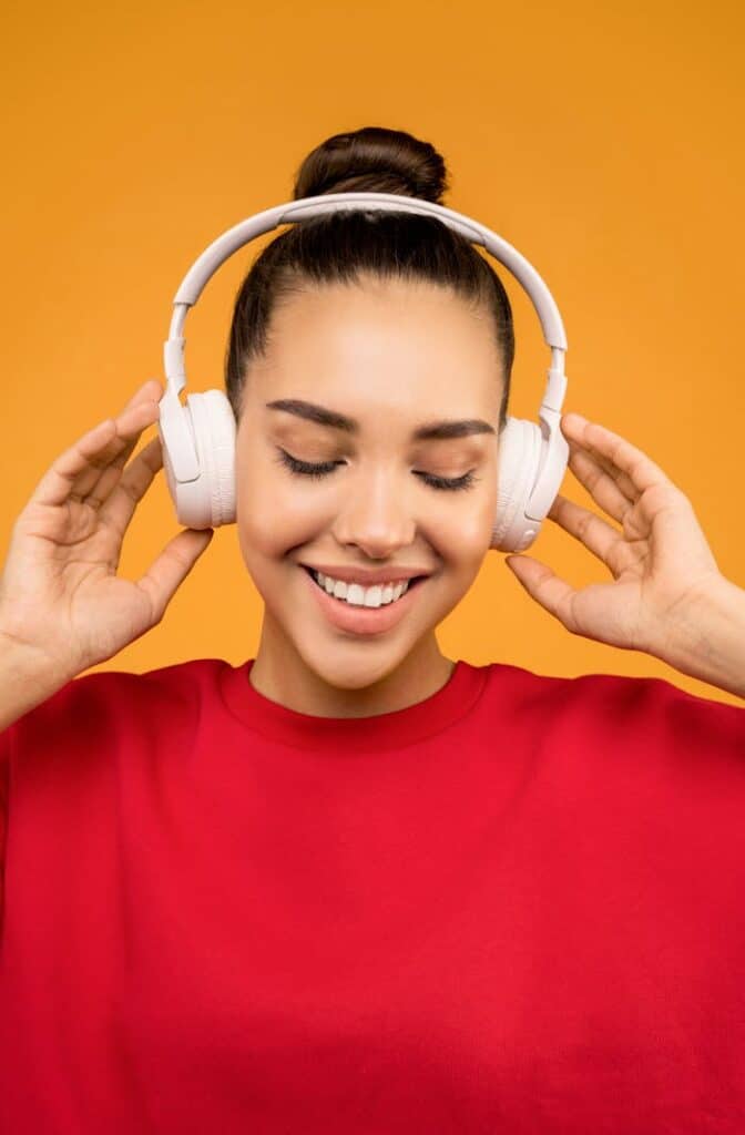 pexels-photo-3756767 Smiling woman in red shirt listening to music on headphones against an orange background.