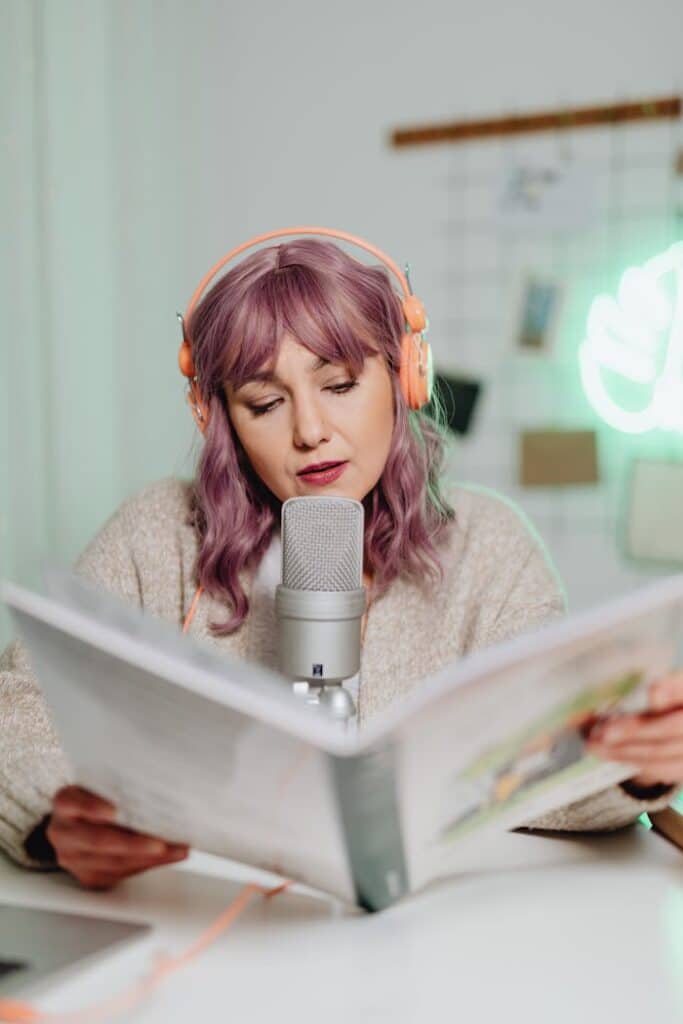 pexels-photo-6919992 Woman podcasting indoors, reading from a book with microphone and headphones.